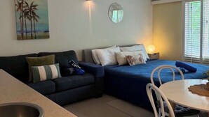 A compact living space with a kitchenette, a dining area, a bed with a blue bedspread, a wall-mounted air conditioner, and a painting of palm trees. - Apart King & Double Sofa Bed Kitchenette Heated Pool WIFI Big TV Walk to Beach (Port Douglas)
