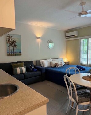 A compact living space with a kitchenette, a dining area, a bed with a blue bedspread, a wall-mounted air conditioner, and a painting of palm trees.