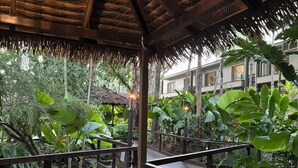 A wooden deck with a thatched roof, surrounded by lush greenery and a view of a building in the background. - Apart King & Double Sofa Bed Kitchenette Heated Pool WIFI Big TV Walk to Beach (Port Douglas)