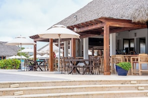 An outdoor seating area with wooden tables and chairs, umbrellas, and a thatched roof structure.