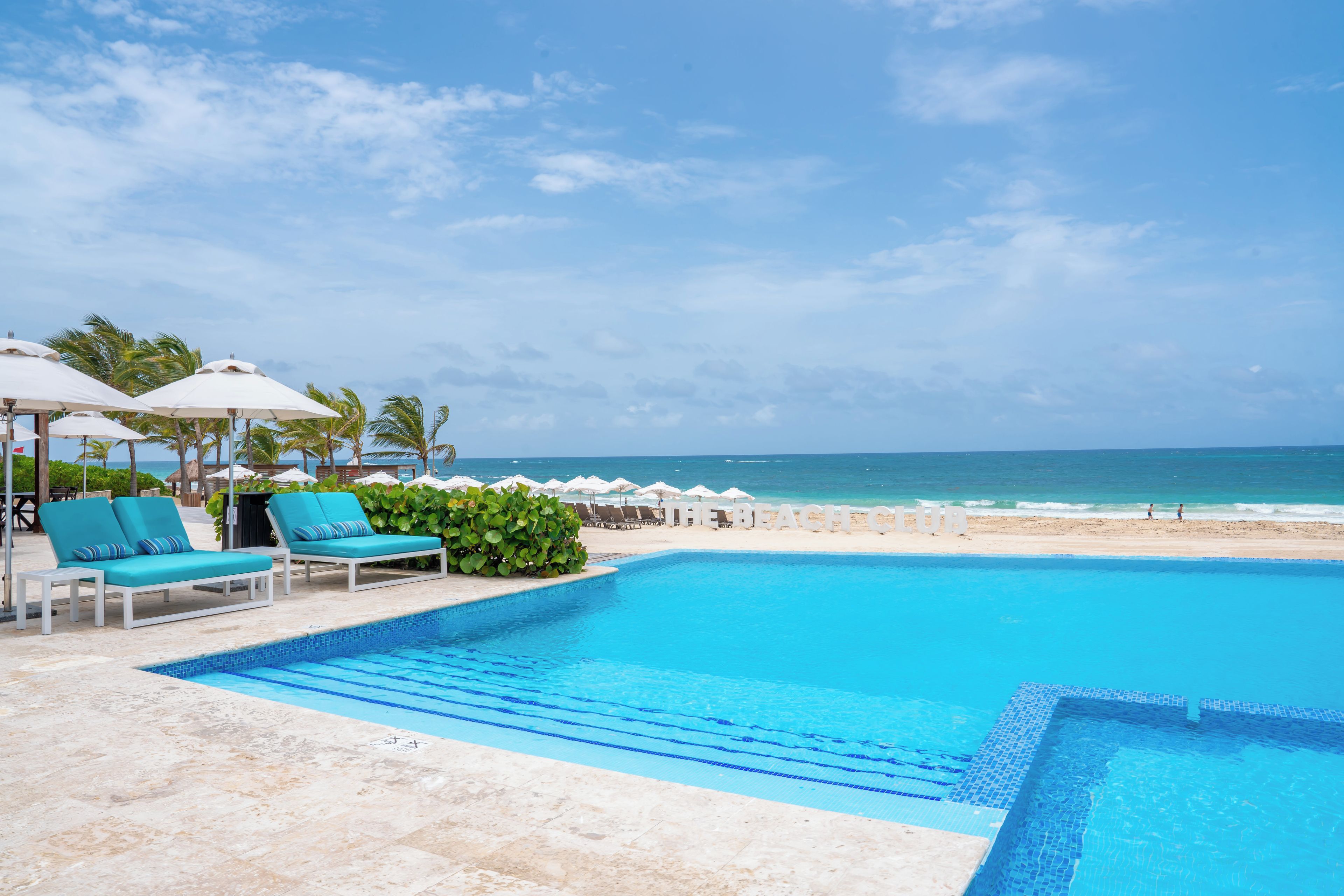 Ein Poolbereich mit Liegestühlen und Sonnenschirmen, Blick auf einen Strand und ein klarer Himmel.