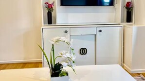 A modern living room with a flat-screen TV mounted on a white wall, a white table with a vase of white flowers, and a cabinet with a lock.