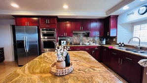 A modern kitchen with stainless steel appliances, dark red cabinets, a granite countertop, and a clock on the wall.