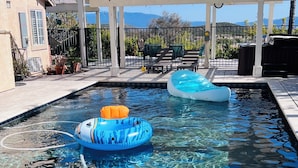 A clear swimming pool with two inflatable rings, a covered patio area with seating, and a view of distant mountains.