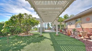 A covered patio area with artificial grass, potted plants, and a view of a house in the background.