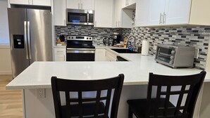 A modern kitchen with white cabinetry, a central island with two black chairs, a stainless steel refrigerator, a microwave, and a toaster oven. - Charming 3-bedroom house in Portland (Portland)