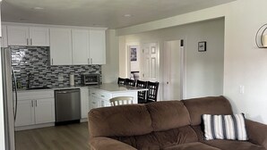 A living room with a brown sofa, a dining area with a table and chairs, a kitchen with white cabinets and a stainless steel refrigerator. - Charming 3-bedroom house in Portland (Portland)
