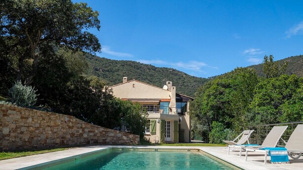 A pool with lounge chairs, a stone wall, and a house with a balcony.