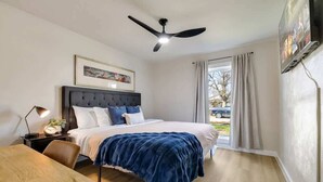 Un dormitorio con cama, escritorio, ventilador de techo, ventana con cortinas y un cuadro en la pared.