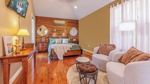 Un dormitorio con cabecera de madera, cama con manta verde, sofá blanco, taburete pequeño y televisor montado en la pared.