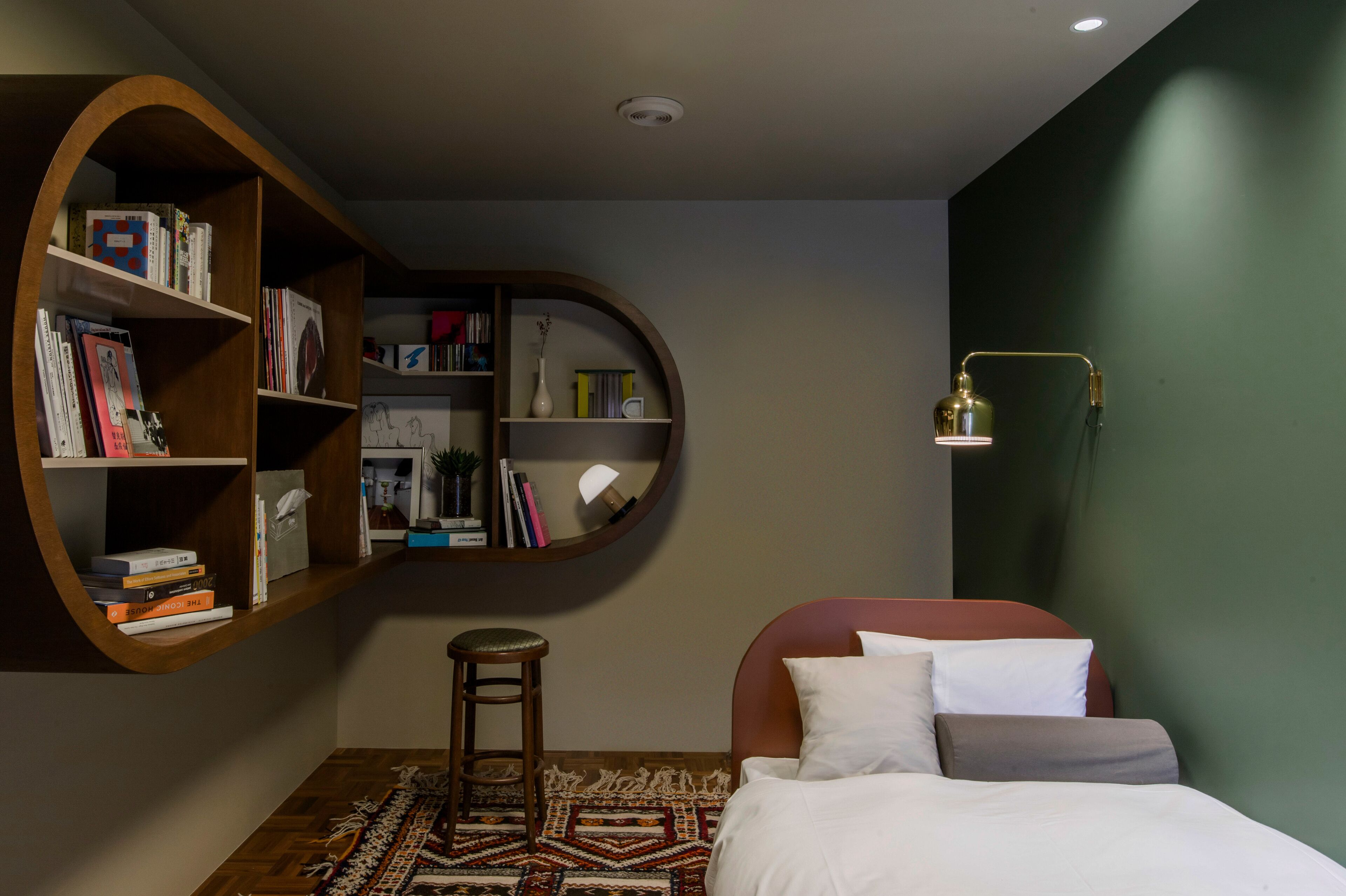 Un dormitorio con cama, estantería, taburete y lámpara.