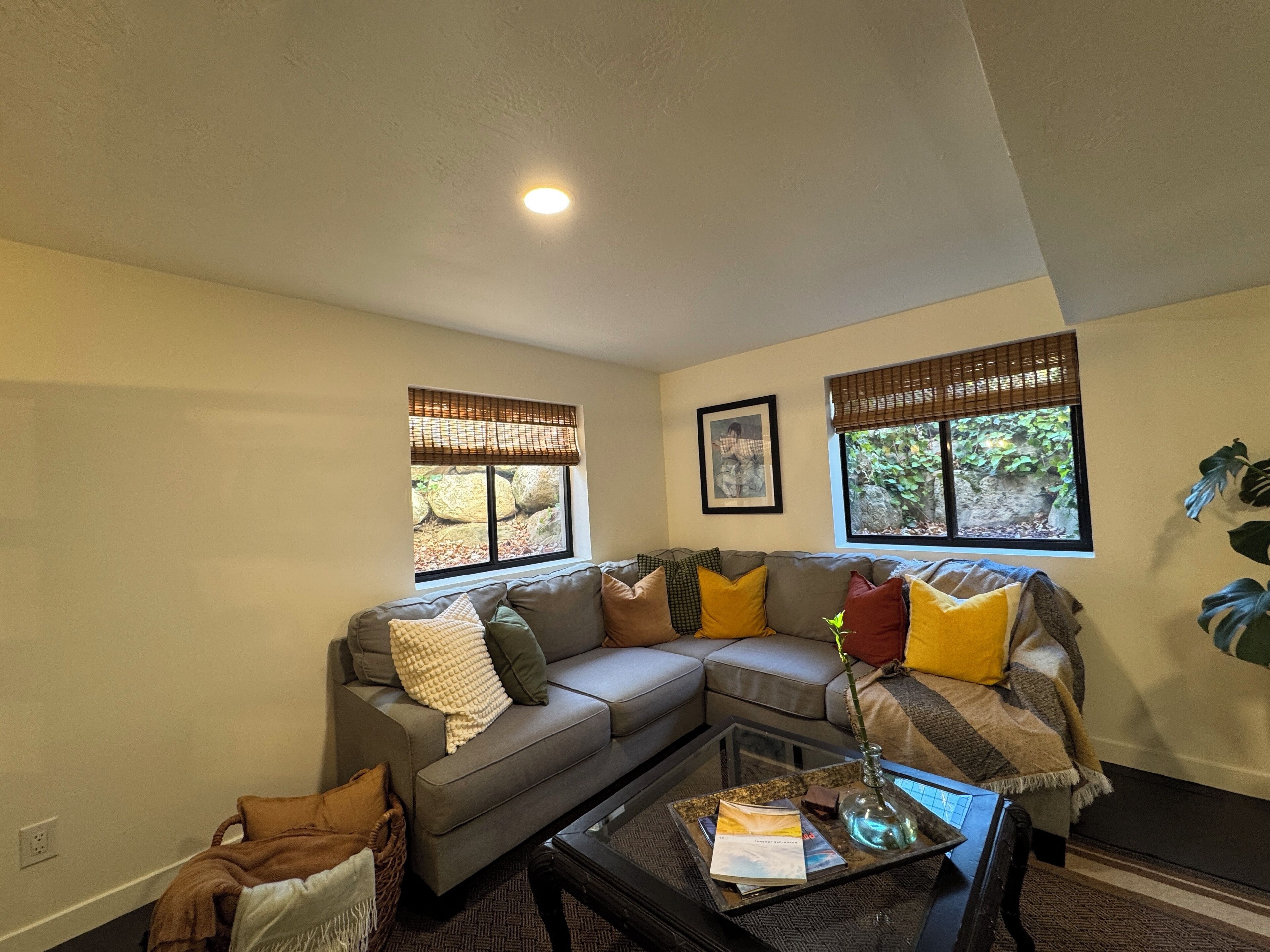 A living room with a gray sectional sofa, a coffee table with magazines and a vase, and two windows with blinds.