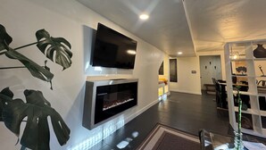 A modern living room with a flat-screen TV mounted on a white wall, a fireplace below it, and large green leaves as wall decor. - Rustic luxe Hideaway-near Beautiful American Fork Canyon! (Highland)