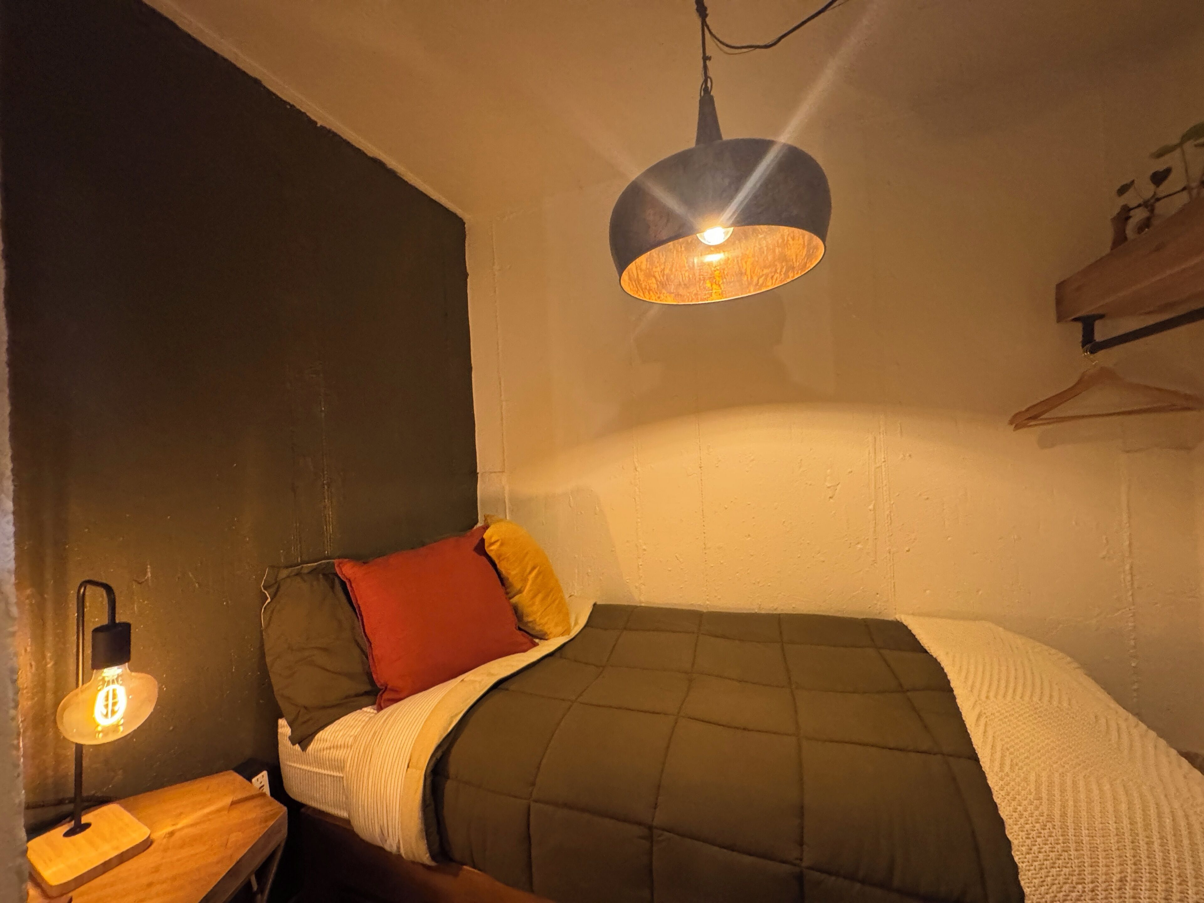A bedroom with a bed, a bedside table, a hanging light fixture, and a wall-mounted shelf.