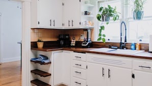 Cocina con armarios blancos, encimera de madera, cafetera, licuadora y ventana con plantas.