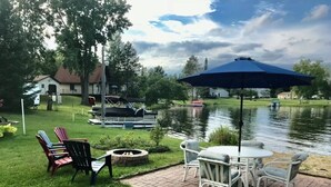 Eine Terrasse mit Gartenmöbeln und einem Feuerkorb, mit Blick auf einen See mit Booten und Häusern im Hintergrund.
