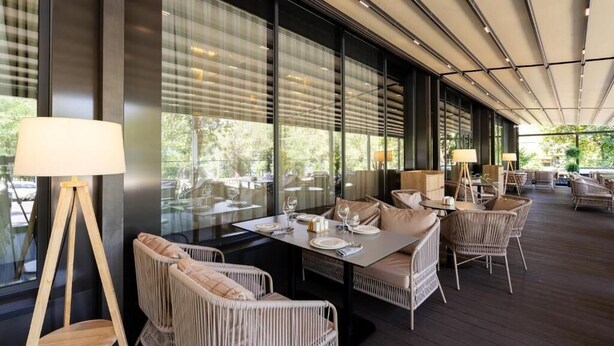 Zona pranzo all'aperto con mobili in vimini, ampie finestre e un soffitto con luci lineari.