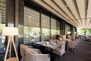 An outdoor dining area with wicker furniture, large windows, and a ceiling with linear lights. - Renion Almaty Hotel (Almaty)