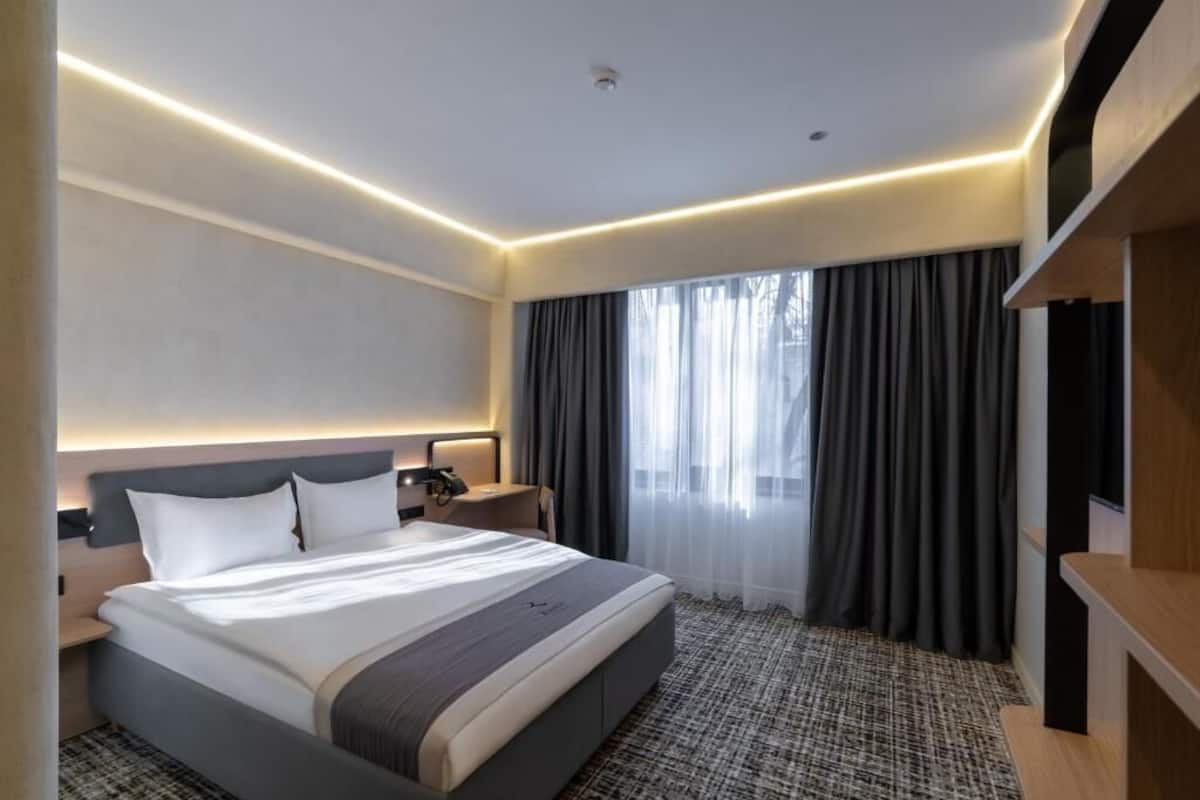 Une chambre d’hôtel moderne avec un grand lit, une table de chevet, une télévision et une fenêtre avec des rideaux.