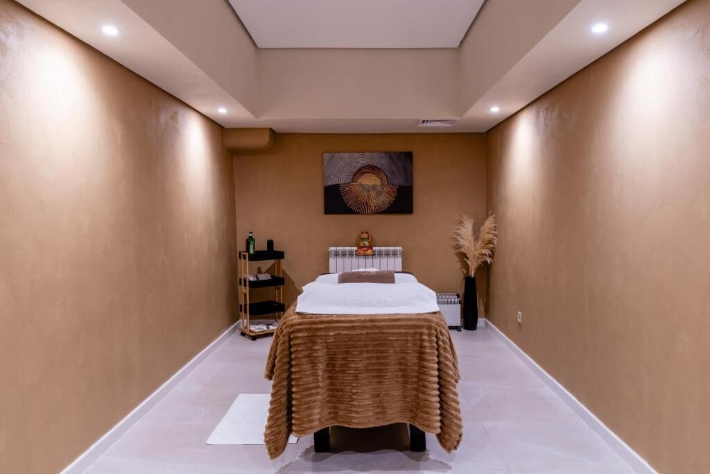 A spa room with a bed, a massage table, a shelf with bottles, and a painting on the wall.