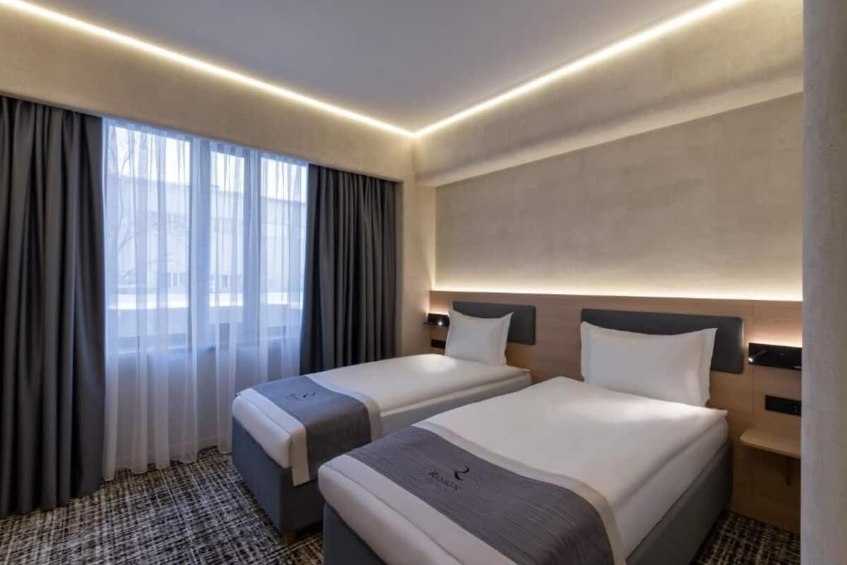 Une chambre d’hôtel avec deux lits, une grande fenêtre avec des rideaux, une tête de lit dotée d’un éclairage intégré et une moquette à motifs.