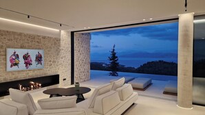 A modern living room with a stone wall, a fireplace, and a large window offering a view of a tree and a lake. - Luxury Villa with Infinity Pool & Sea Views in Northeast Corfu (kassiopi)