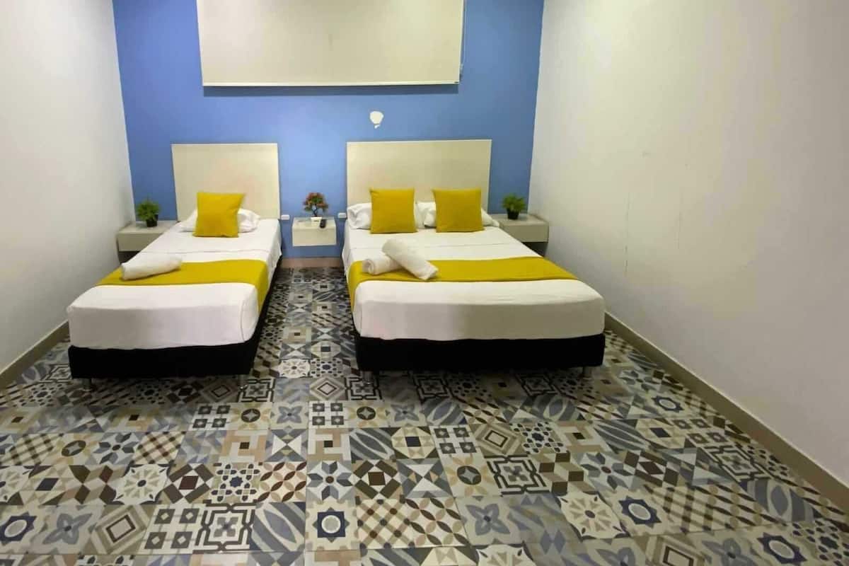 A hotel room with two beds, each with a white mattress and yellow pillows, a patterned floor, and an air conditioning unit on the wall.