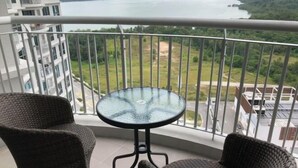 A balcony with wicker chairs and a glass table, overlooking a body of water and greenery. - Teega Residence Waterfront Living (Iskandar Puteri)
