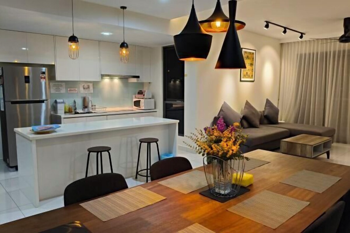 A modern kitchen with a central island, bar stools, pendant lighting, a dining table with placemats, and a living area with a sofa and a coffee table.