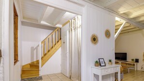A spacious room with a wooden staircase, white walls, and a tiled floor. There is a TV, a small table with decorative items, and a sunburst mirror on the wall.