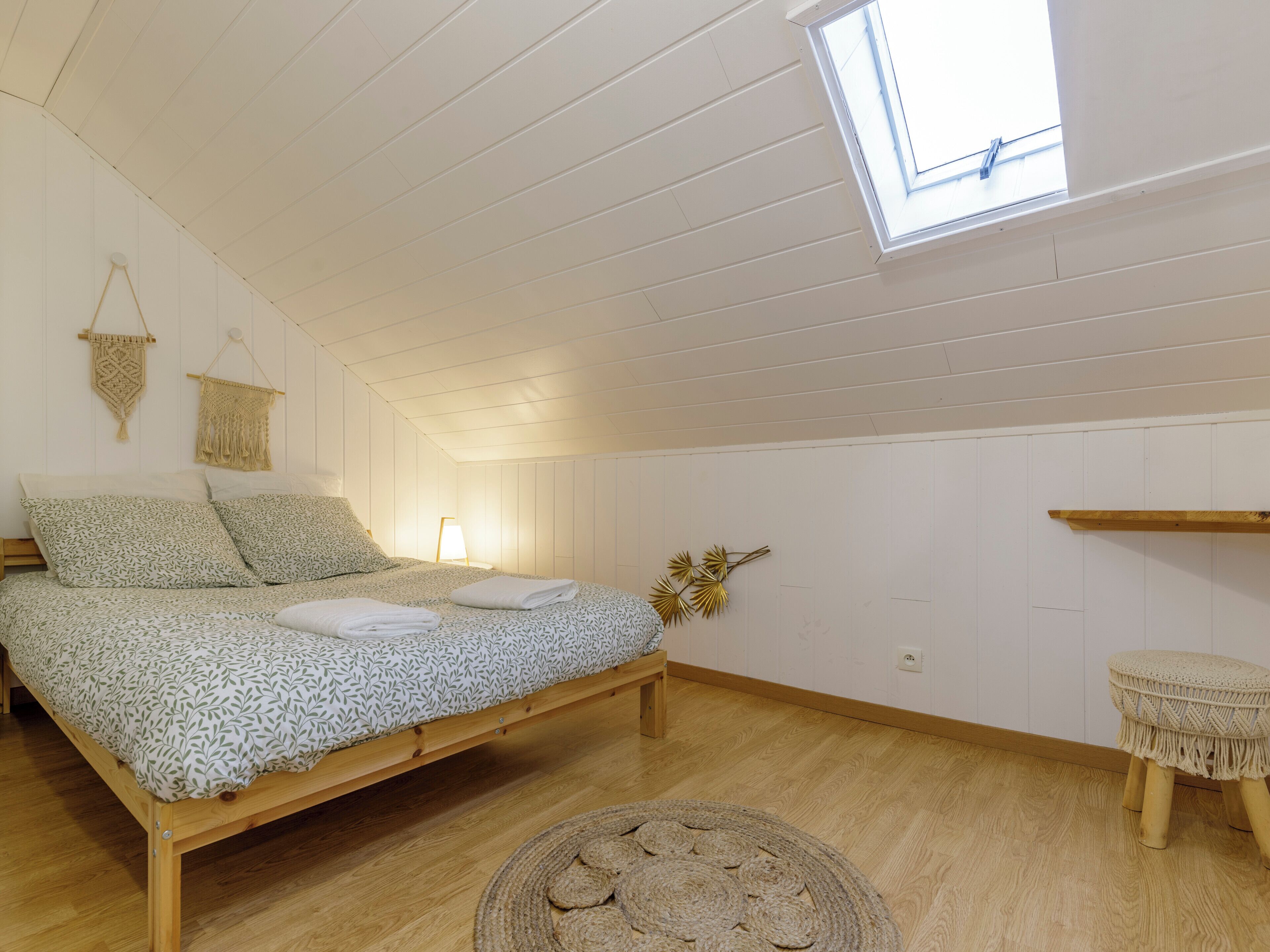 Ein Schlafzimmer mit einem Bett, einem kleinen Hocker, einem Wandregal und einem Dachfenster.