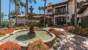 A courtyard with a fountain, palm trees, and a building with a balcony. - Luxury 2BR/2BA Smart Home in Monterey Country Club – Palm Desert, CA (Palm Desert)