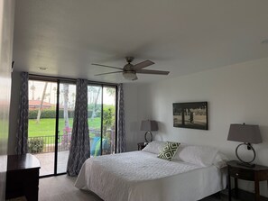 Une chambre à coucher avec un lit, deux tables de chevet avec des lampes, un ventilateur de plafond, une grande fenêtre avec des rideaux et un tableau encadré au mur.