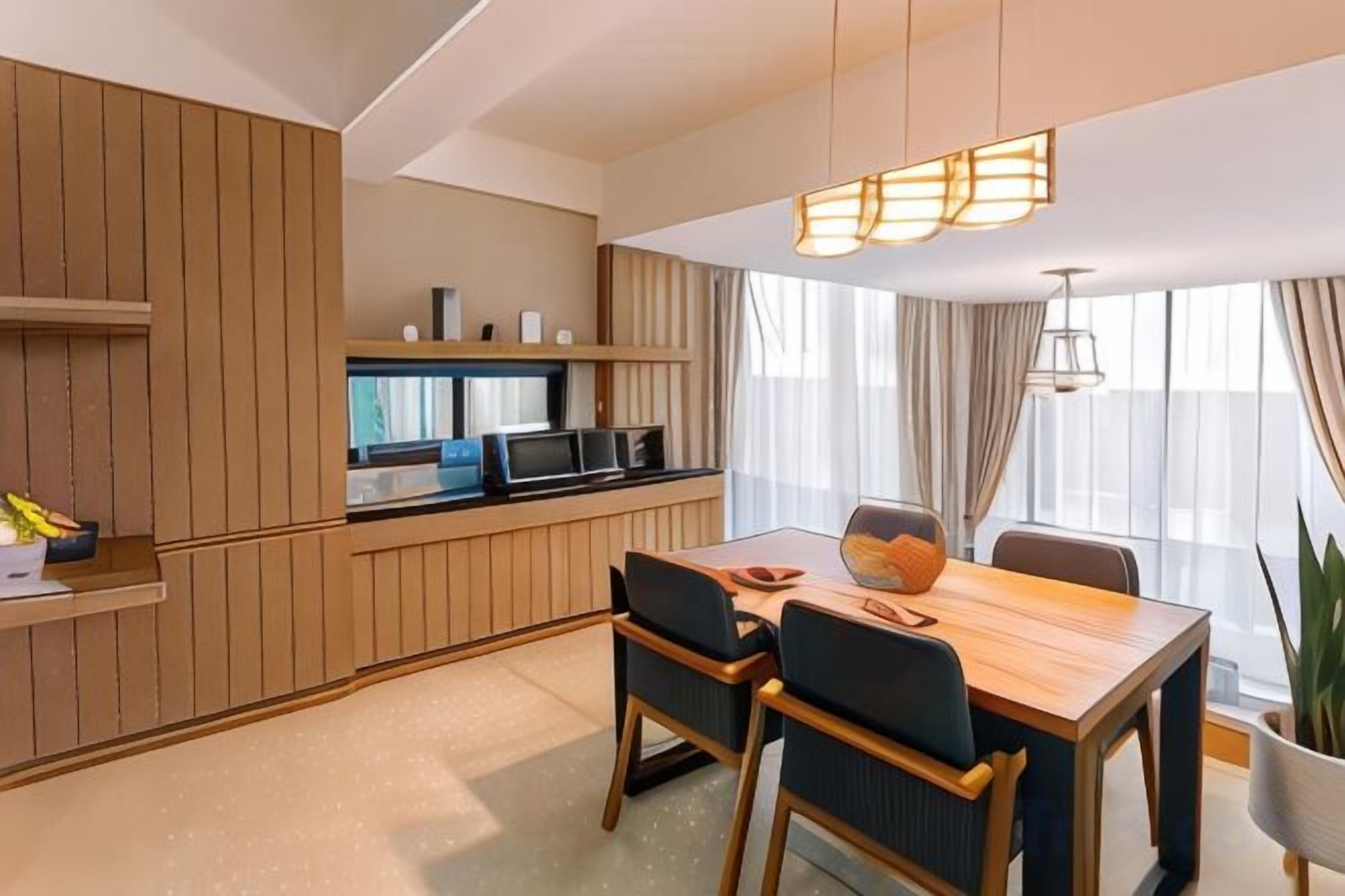 A modern hotel room with a wooden dining table, two chairs, a kitchen area with a microwave, and a large window with curtains.