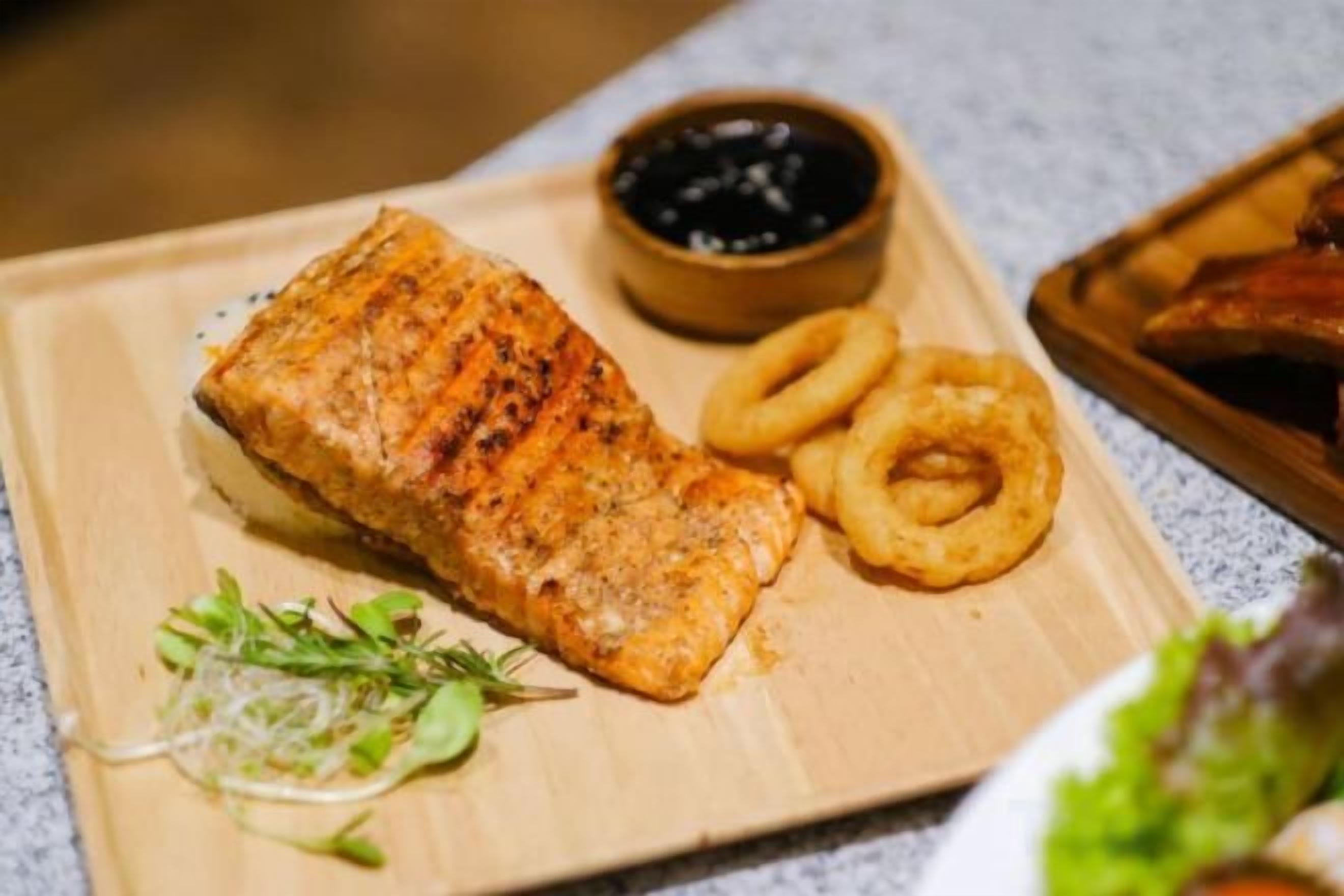 A wooden cutting board with grilled salmon, onion rings, and a small bowl of sauce.