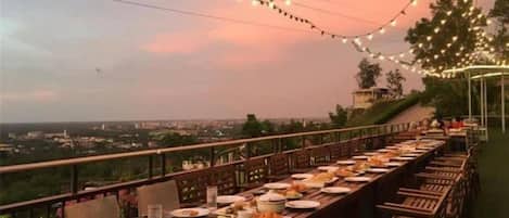 An outdoor dining area with tables and chairs, string lights, and a view of the city.