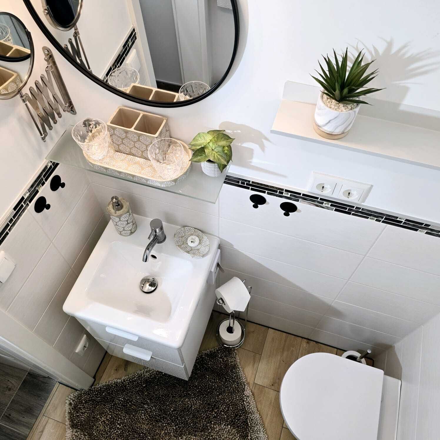 A modern bathroom with a white sink, mirror, and toilet. There is a small plant on a shelf and a decorative tray with glassware.