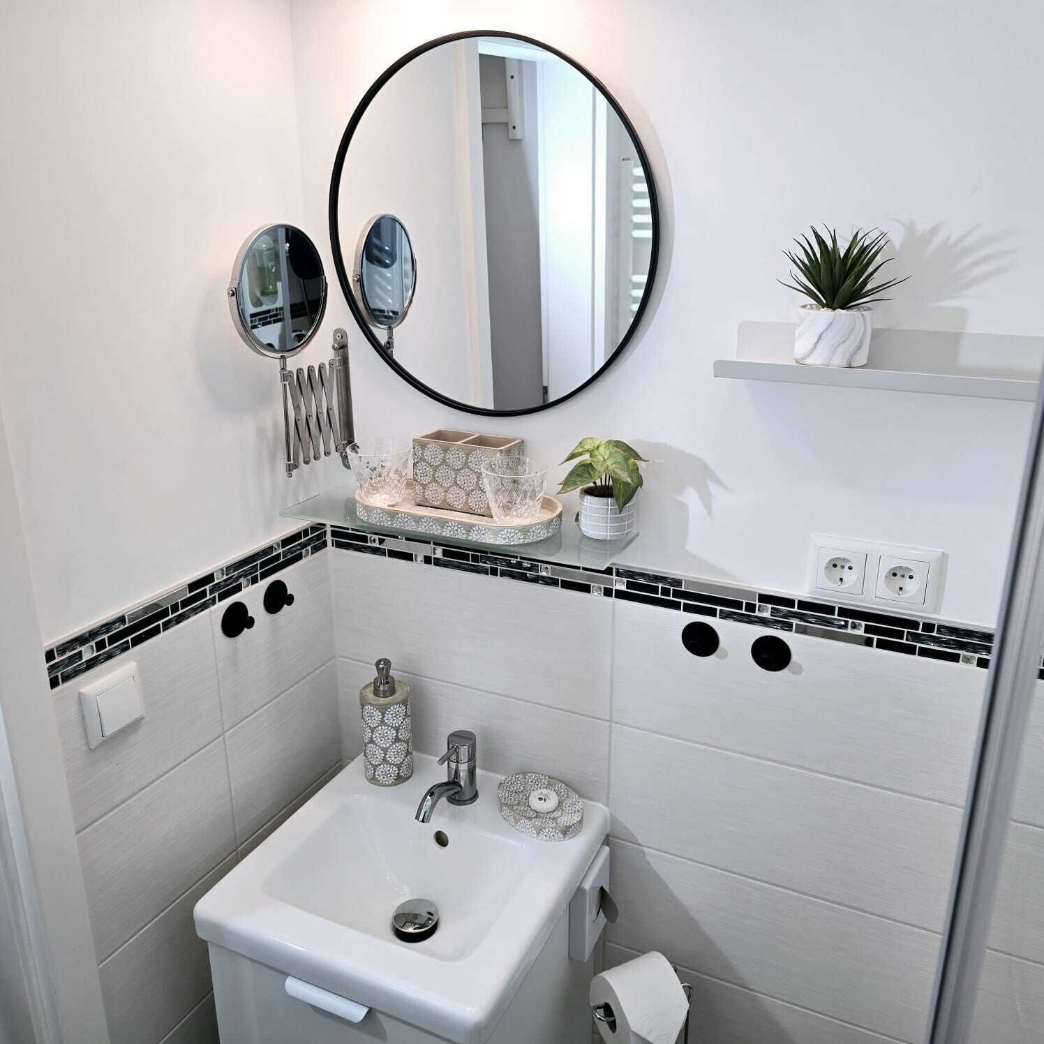 Ein Badezimmer mit einem weißen Waschbecken, zwei Spiegeln, einer kleinen Ablagefläche mit einer Pflanze und einer Toilette.