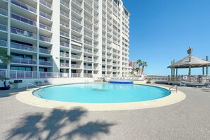 Zona de piscina de hotel con una piscina grande, sillas de descanso y un edificio con varios balcones.