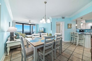 A dining area with a view of the ocean, a ceiling fan, and a kitchen with a blue backsplash.