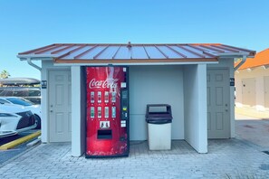 A Coca-Cola vending machine in a small structure with a metal roof, a trash bin, and a parking lot with cars. - Summer House 905A | Gulf Front | Resort Amenities | My Beach Getaways (Orange Beach)