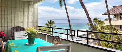 Uma varanda com mesa e cadeiras, com vista para uma praia com palmeiras e o oceano.