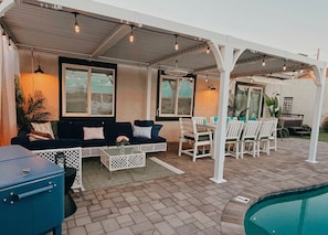 An outdoor patio area with a pool, a covered seating area with a sofa and chairs, and a grill.