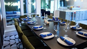 A dining area with a long table set for a meal, a view of the outdoors through large windows, and a modern kitchen in the background. - Lunar: Old Town Luxury• Heated Pool• 2 Kings• Gameroom• Hottub• Karaoke• Privacy (Scottsdale)