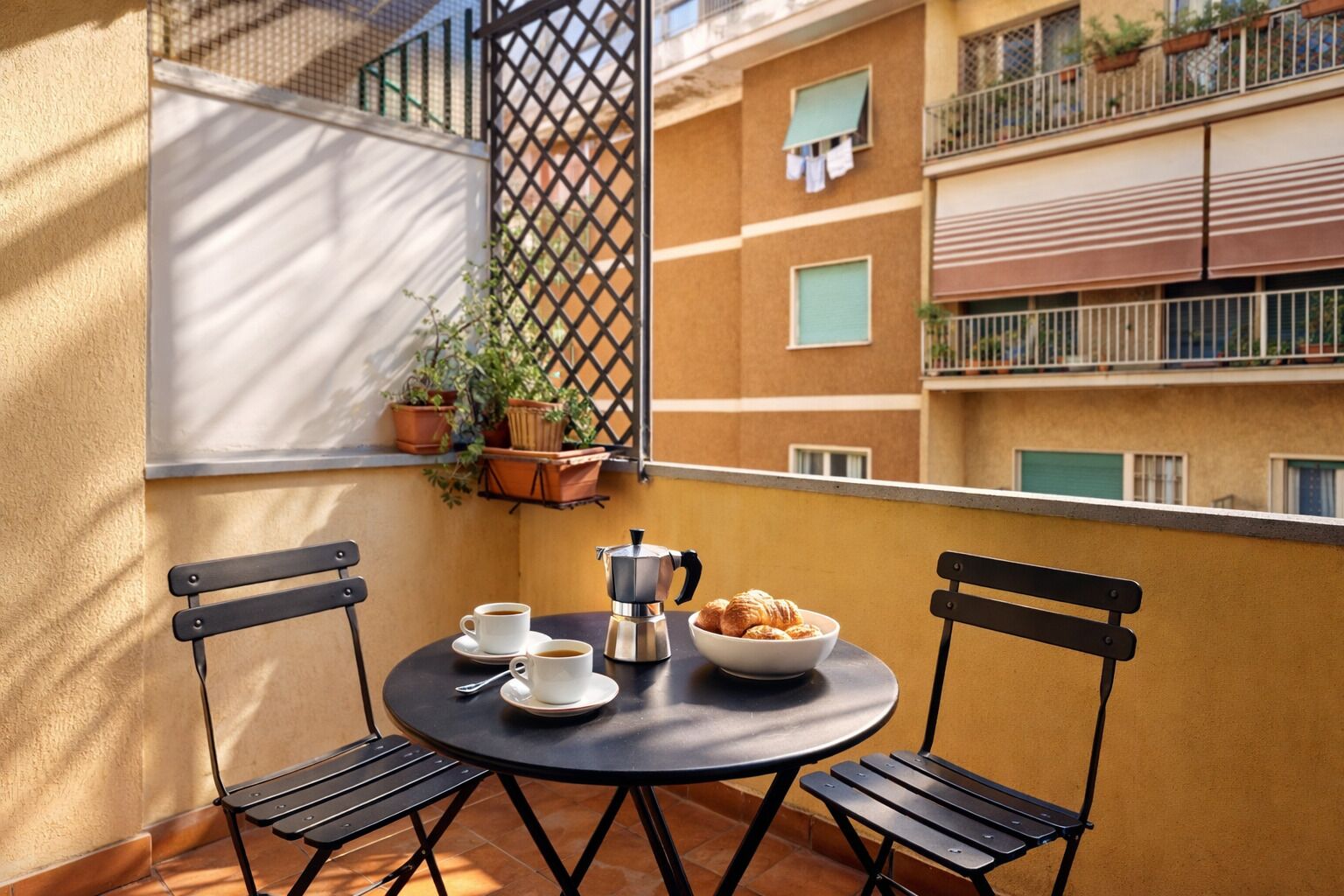 Ein Balkon mit zwei Stühlen, einem Tisch, einer Kaffeemaschine, Tassen und einer Schüssel Croissants.