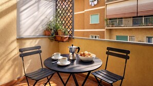 En balkong med två stolar, ett bord, en kaffebryggare, koppar och en skål med croissanter.