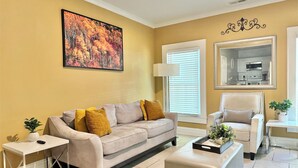 A living room with a beige sofa, a white side table, a small potted plant, a framed picture on the wall, and a kitchen area visible through an open door.