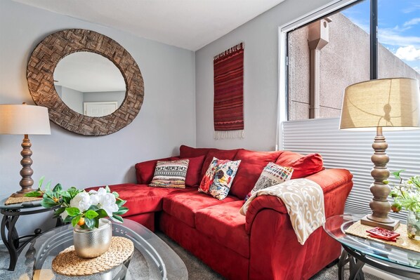 A red sofa with patterned cushions, a round mirror, a red tapestry, and a lamp on a glass table.
