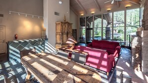 A spacious room with a wooden ceiling, a large window with a view of greenery, a red sofa, a wooden table, and a vintage lamp.