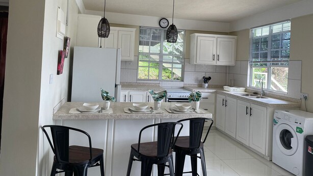 A modern kitchen with white cabinets, a built-in oven, a sink, and a washing machine. There are two black chairs and a small island with a sink and a clock. - Providence,where nature meets home, every path leads to peace.
Strong WIFI, AC. (Mahaut)